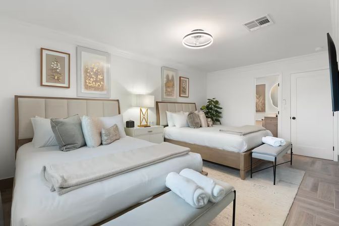 Bright modern guest bedroom with two queen beds, neutral linens, bedside lamp, rolled spa towels on benches, framed botanical art and a potted plant.