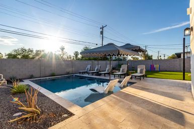 Sunny southwestern backyard pool at golden hour with two in-pool loungers, row of chaise lounges under scalloped umbrella, stone tile patio, artificial turf and desert plants