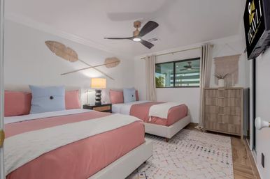 Bright modern guest bedroom with two twin beds in pink-and-white bedding, woven wall art, black ceiling fan, nightstand with lamp, wood dresser, patterned rug, and a window letting in natural light.