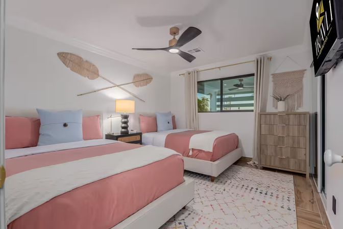 Bright modern guest bedroom with two twin beds in pink-and-white bedding, woven wall art, black ceiling fan, nightstand with lamp, wood dresser, patterned rug, and a window letting in natural light.