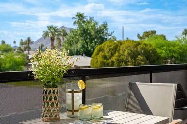Sunlit balcony with mountain and palm trees in the distance, cheery daisies in a woven vase on a patio table beside a pitcher and two lemon-water glasses — bright, relaxed outdoor scene.