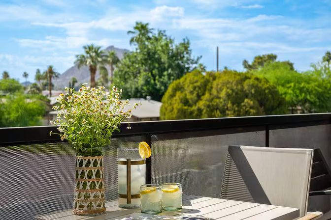 Sunlit balcony with mountain and palm trees in the distance, cheery daisies in a woven vase on a patio table beside a pitcher and two lemon-water glasses — bright, relaxed outdoor scene.