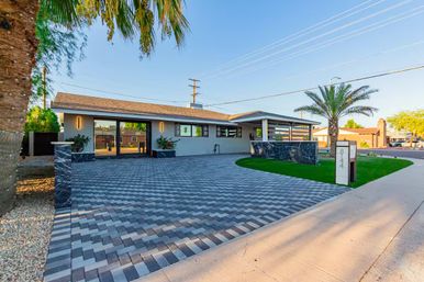 Sunlit modern ranch-style home in a sunny desert suburb with chevron-pattern gray paver driveway, low dark stone walls, trimmed green turf and palm trees