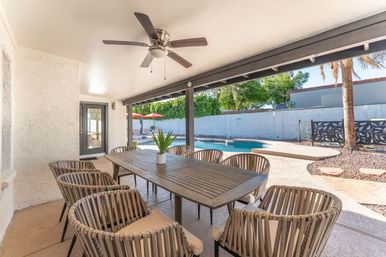 Covered backyard patio with wooden outdoor dining table and eight woven chairs, ceiling fan overhead, overlooking a sunny swimming pool, palm tree and patio umbrellas.