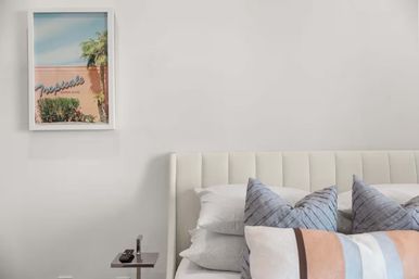 Bright modern bedroom with white upholstered headboard, layered decorative pillows, small side table with remote, and framed pastel tropical art featuring a palm tree on a pale wall.
