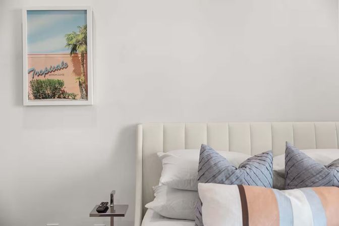 Bright modern bedroom with white upholstered headboard, layered decorative pillows, small side table with remote, and framed pastel tropical art featuring a palm tree on a pale wall.