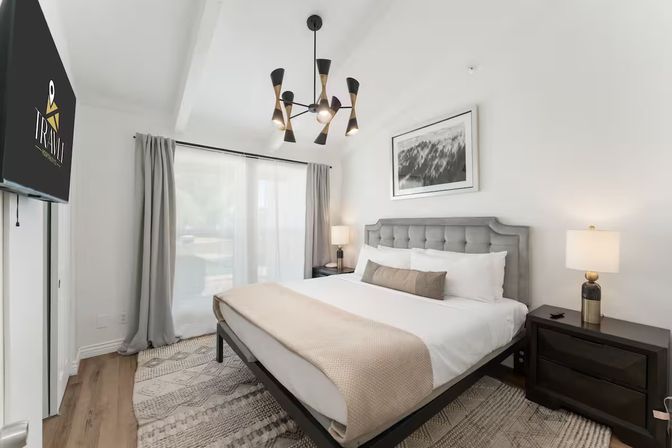 Bright modern bedroom with king bed and tufted gray headboard, beige throw and lumbar pillow, mid-century chandelier, bedside lamps, wall-mounted TV, sheer curtains over sliding glass door, hardwood floor and patterned rug.