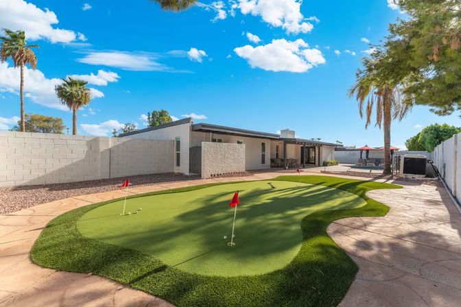 Sunny backyard with synthetic putting green and three red flags, adjacent patio with seating and red umbrellas, single-story modern house, palm trees and bright blue sky.