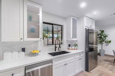 Bright modern white kitchen with shaker cabinets, marble-look countertops, black undermount sink and gooseneck faucet, stainless steel refrigerator and dishwasher, wood-look tile floor, potted plant and bowl of lemons