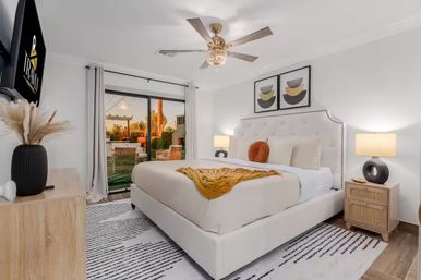 Bright modern bedroom with a tufted upholstered bed, mustard throw and round rust pillow, wood nightstands with sculptural lamps, ceiling fan, wall art and flat-screen TV, sliding glass doors opening to a furnished patio at sunset.