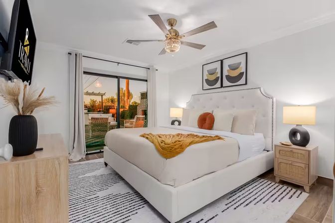 Bright modern bedroom with a tufted upholstered bed, mustard throw and round rust pillow, wood nightstands with sculptural lamps, ceiling fan, wall art and flat-screen TV, sliding glass doors opening to a furnished patio at sunset.