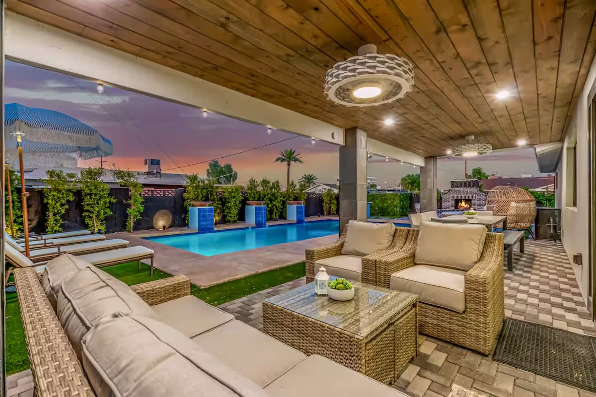 Modern covered backyard patio at sunset with wicker sofa and armchairs, glass-top coffee table, illuminated rectangular pool, string lights and outdoor fireplace