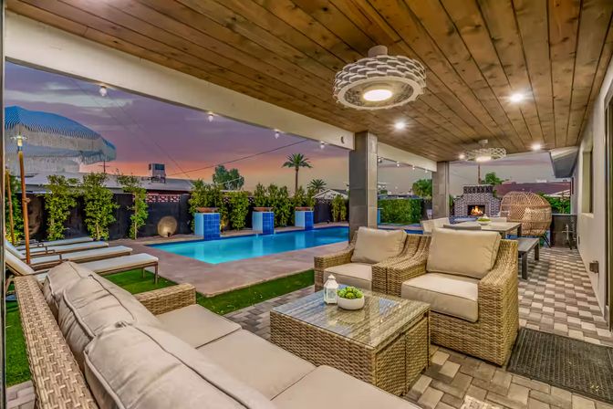 Modern covered backyard patio at sunset with wicker sofa and armchairs, glass-top coffee table, illuminated rectangular pool, string lights and outdoor fireplace