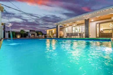 Dusk view of a luxury backyard swimming pool with turquoise water, illuminated modern patio, outdoor dining table, lounge seating and sunset sky