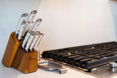 Sleek stainless-steel–handled knife set in a wooden block on a white kitchen countertop beside a black gas stovetop, modern home kitchen prep area.