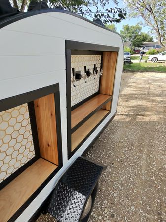 Mobile bar trailer with four beer taps, honeycomb tile backsplash and wooden shelves, parked on a gravel driveway under trees
