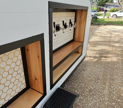Mobile bar trailer with four beer taps, honeycomb tile backsplash and wooden shelves, parked on a gravel driveway under trees