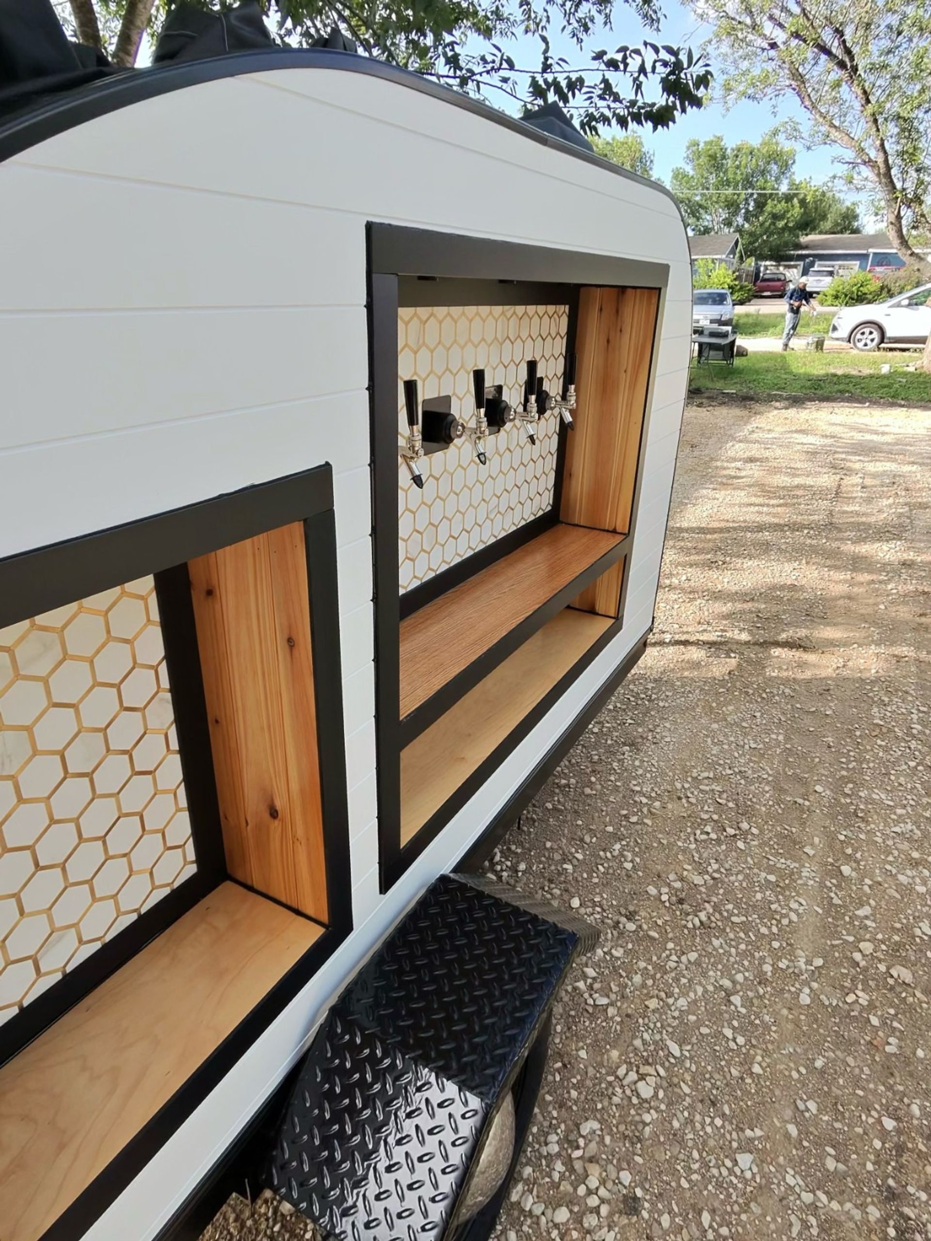 Mobile bar trailer with four beer taps, honeycomb tile backsplash and wooden shelves, parked on a gravel driveway under trees
