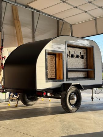 Compact black-and-white teardrop trailer converted into a mobile bar, with an open serving window, wood shelving, honeycomb tile backsplash and four beer taps, parked inside a sunlit garage on a single-axle trailer.