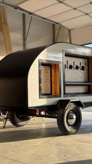 Compact black-and-white teardrop trailer converted into a mobile bar, with an open serving window, wood shelving, honeycomb tile backsplash and four beer taps, parked inside a sunlit garage on a single-axle trailer.