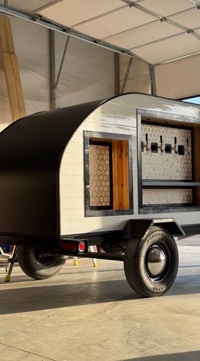 Compact black-and-white teardrop trailer converted into a mobile bar, with an open serving window, wood shelving, honeycomb tile backsplash and four beer taps, parked inside a sunlit garage on a single-axle trailer.
