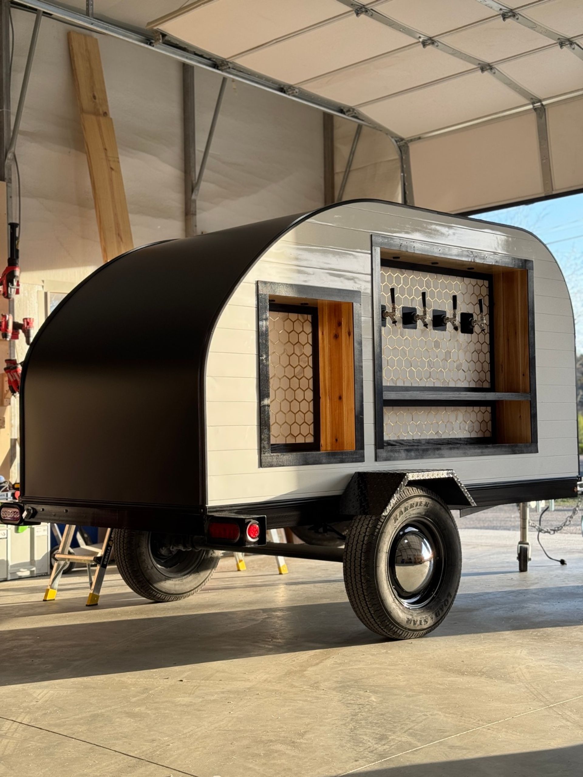Compact black-and-white teardrop trailer converted into a mobile bar, with an open serving window, wood shelving, honeycomb tile backsplash and four beer taps, parked inside a sunlit garage on a single-axle trailer.