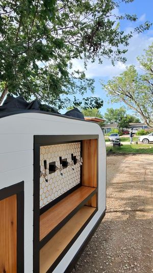 Mobile white trailer bar with four beer taps, honeycomb tile backsplash and wood shelves, parked on a gravel driveway under trees in a suburban backyard.