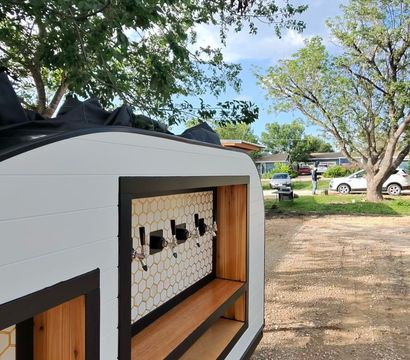 Mobile white trailer bar with four beer taps, honeycomb tile backsplash and wood shelves, parked on a gravel driveway under trees in a suburban backyard.