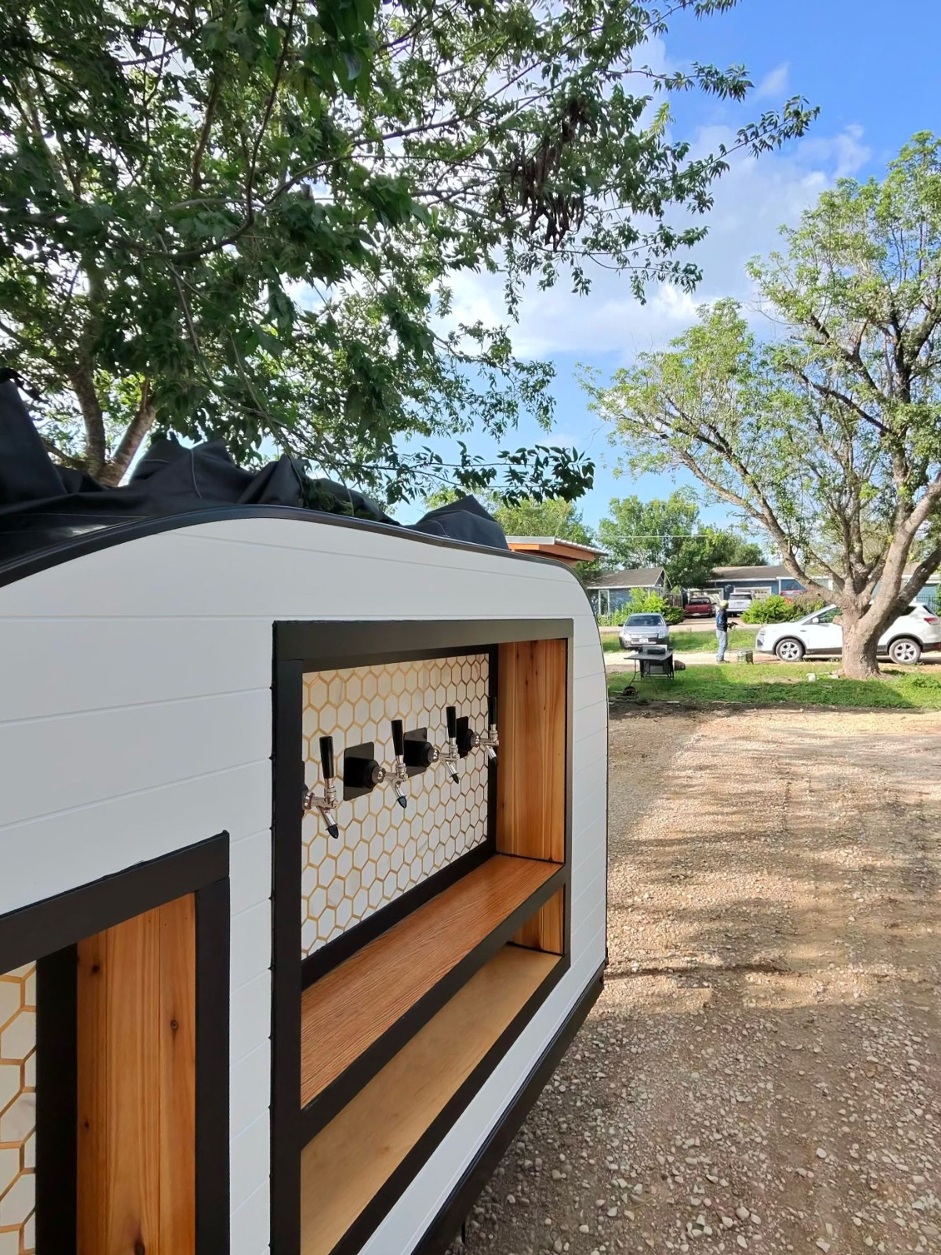 Mobile white trailer bar with four beer taps, honeycomb tile backsplash and wood shelves, parked on a gravel driveway under trees in a suburban backyard.