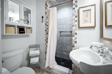 Bright small bathroom with patterned tile-framed shower, gray marble shower walls and black pebble floor, white pedestal sink with chrome faucet, toilet, mirrored wall cabinet with wicker baskets and folded towels.