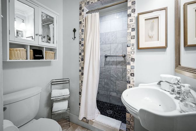 Bright small bathroom with patterned tile-framed shower, gray marble shower walls and black pebble floor, white pedestal sink with chrome faucet, toilet, mirrored wall cabinet with wicker baskets and folded towels.