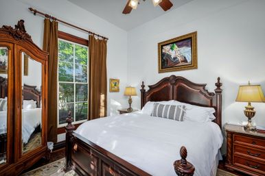 Sunlit Victorian-style bedroom with ornate carved dark wood bed and matching armoire, crisp white linens, bedside lamps, framed artwork, and a tall window with brown curtains overlooking a leafy yard.