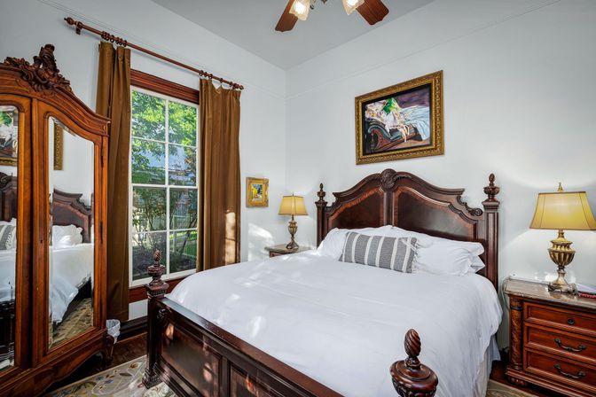 Sunlit Victorian-style bedroom with ornate carved dark wood bed and matching armoire, crisp white linens, bedside lamps, framed artwork, and a tall window with brown curtains overlooking a leafy yard.