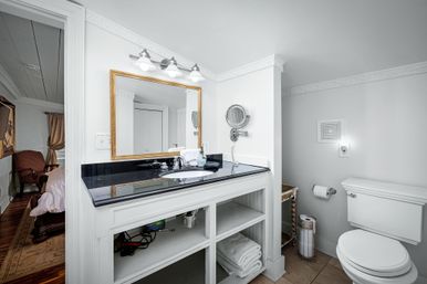 Bright white bathroom with black granite vanity, gold-framed mirror and triple lights, open shelving with folded towels, wall-mounted magnifying mirror, toilet and doorway to bedroom.