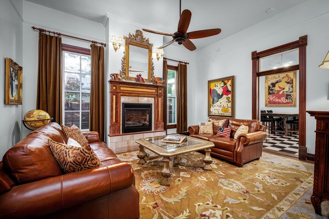 Victorian-style living room with brown leather sofas, ornate wood fireplace topped by a gilded mirror, patterned area rug, glass-top coffee table, ceiling fan, vintage posters and a globe.