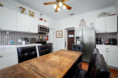 White modern farmhouse kitchen with wooden dining table, black leather chairs, stainless steel refrigerator and microwave, geometric tile backsplash and ceiling fan