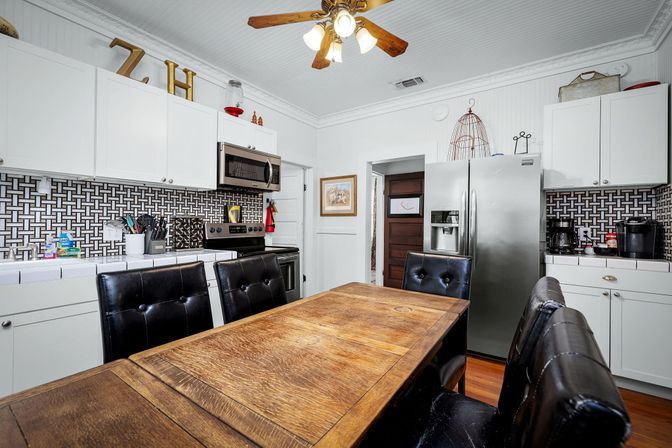 White modern farmhouse kitchen with wooden dining table, black leather chairs, stainless steel refrigerator and microwave, geometric tile backsplash and ceiling fan