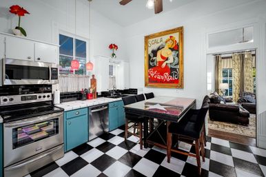 Bright retro kitchen with black-and-white checkered floor, teal lower cabinets, stainless steel appliances, glass-topped island with stools, vintage framed poster and view into a cozy living room.