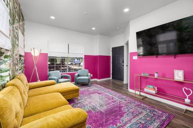 Vibrant modern living room with mustard-yellow sectional, fuchsia half-wall paint, tropical leaf wallpaper, teal armchairs, purple patterned rug, clear acrylic coffee table and wall-mounted TV.