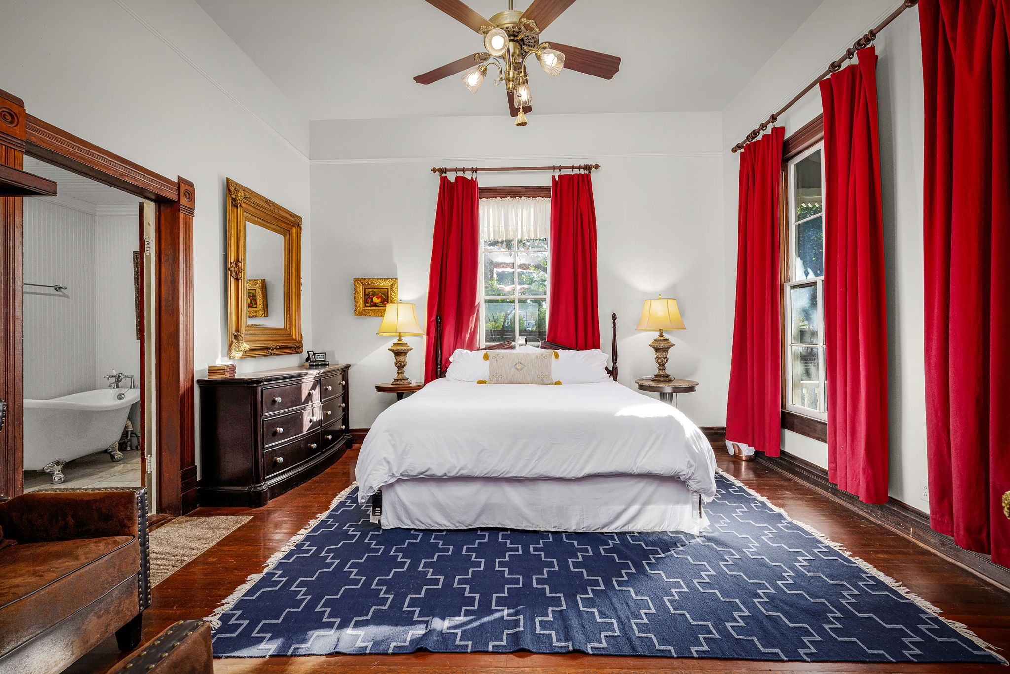 Bright Victorian-style bedroom with a white bed on a navy geometric rug, bold red curtains at tall windows, dark wood furniture and lamps, ceiling fan overhead and a clawfoot bathtub visible through an open doorway.