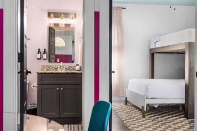 Bright modern hotel-style suite interior showing a granite-top bathroom vanity with mirror and wall-mounted soap dispensers beside a bedroom with a white bunk bed, patterned rug, soft pink curtains and a teal chair.