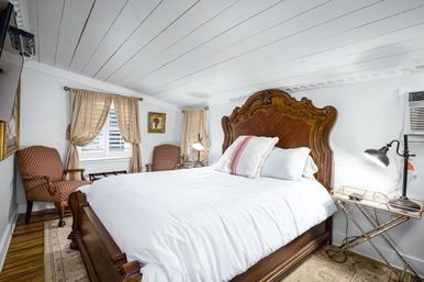 Cozy bright attic bedroom with carved wooden headboard, crisp white bedding, beige curtains over shuttered window, patterned armchairs, bedside lamp and wall air conditioner.
