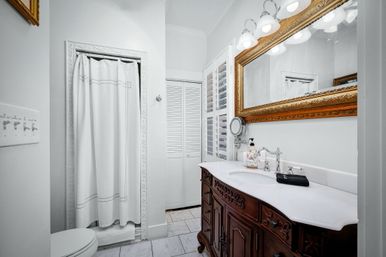 Bright white residential bathroom with carved dark-wood vanity and marble countertop, gold-framed mirror, chrome faucets, white shower curtain, louvered shutters and tile floor