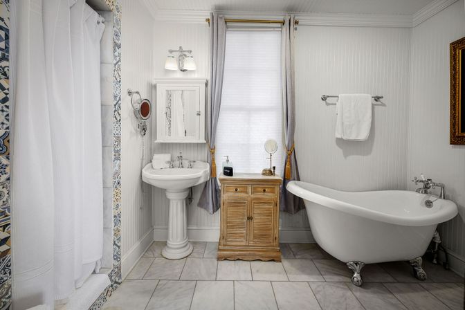 Charming bright bathroom with a white clawfoot tub, pedestal sink, wooden cabinet, marble tile floor, patterned shower tile and a curtained window.