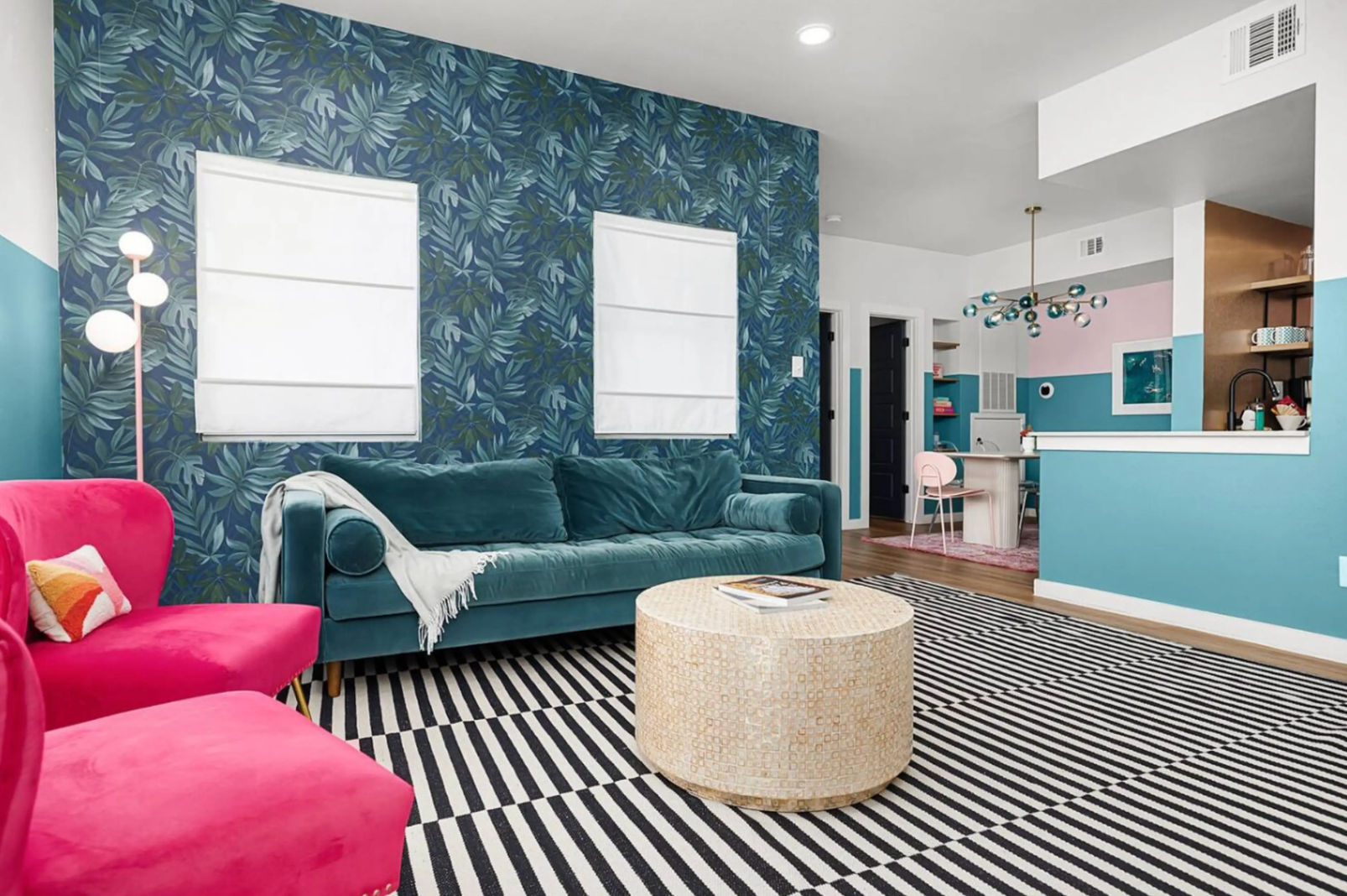 Bright eclectic living room with teal velvet sofa, hot pink armchairs, blue tropical leaf accent wall, black-and-white striped rug, round woven coffee table, and open-plan kitchen with teal and pink accents and a modern chandelier.