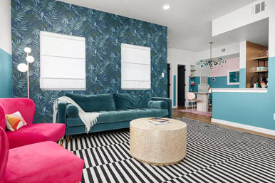 Bright eclectic living room with teal velvet sofa, hot pink armchairs, blue tropical leaf accent wall, black-and-white striped rug, round woven coffee table, and open-plan kitchen with teal and pink accents and a modern chandelier.