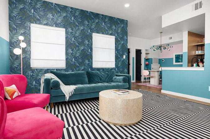 Bright eclectic living room with teal velvet sofa, hot pink armchairs, blue tropical leaf accent wall, black-and-white striped rug, round woven coffee table, and open-plan kitchen with teal and pink accents and a modern chandelier.