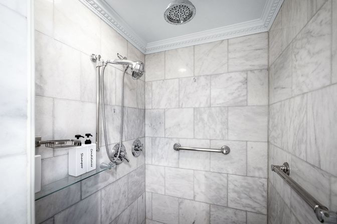 Spa-like marble-tiled shower with chrome rainfall and handheld showerheads, stainless grab bars, and a glass shelf holding labeled shampoo and conditioner bottles.