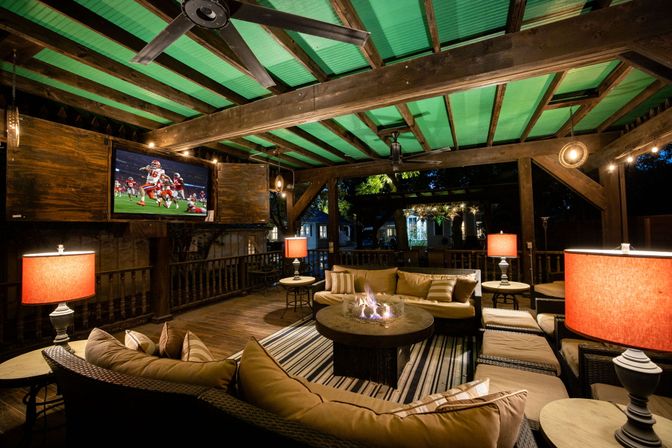 Cozy covered backyard patio at night with wooden beams, sectional sofa around a glowing fire pit, red table lamps, wall-mounted TV showing football, ceiling fans and string lights.