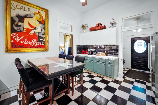 Eclectic bright kitchen with black-and-white checkerboard floor, teal lower cabinets and white uppers, black subway tile backsplash, glass-top breakfast bar with black stools, oversized vintage red-and-gold poster on the wall, and a black door with round porthole window.